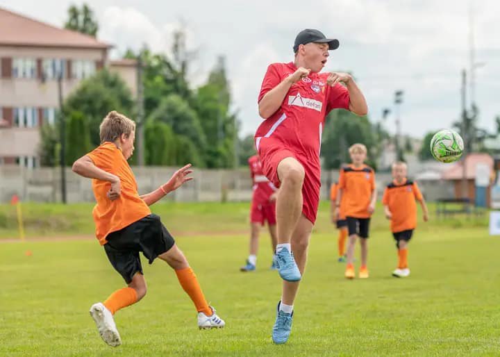 Moment z meczu piłki nożnej, w którym widać dwóch zawodników. Jeden z nich, ubrany w czerwoną koszulkę, przygotowuje się do uderzenia piłki prawą nogą. Drugi zawodnik, w pomarańczowej koszulce, dobiega do pierwszego zawodnika. W tle widać innych zawodników