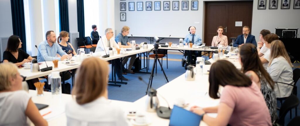 Sala konferencyjna, w której odbywa się spotkanie. Osoby siedzą wokół dużego kwadratowego stołu, na którym leżą dokumenty, mikrofony oraz dzbanki z wodą i kawą.