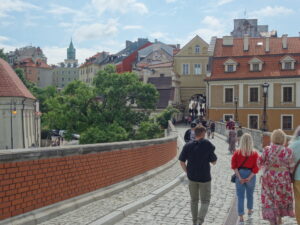 Malowniczy widok Lublina z brukowaną ulicą otoczoną kolorowymi, europejskimi budynkami. Po lewej stronie ceglana ściana mostu, a ludzie spacerują w kierunku rynku.