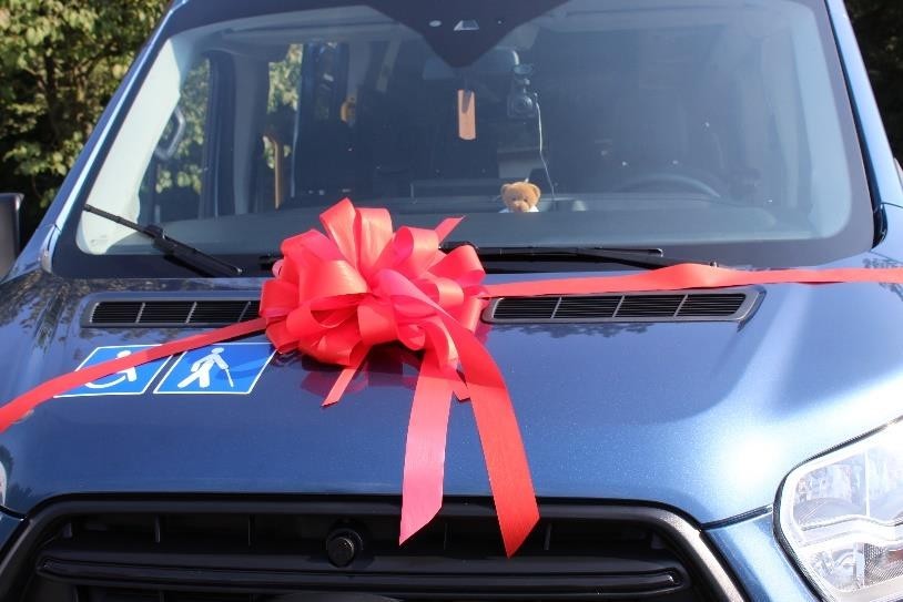 Przód ciemnoniebieskiego busa, na którego masce znajduje się duża czerwona kokarda