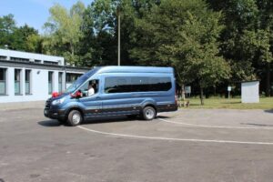 Niebieski bus zaparkowany na asfaltowej powierzchni z białymi liniami parkingowymi. Na przodzie pojazdu znajduje się czerwona wstążka. W tle widać drzewa oraz budynek w kolorze białym z dużymi oknami.