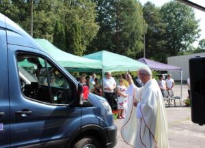 Plenerowe zgromadzenie z kilkoma osobami stojącymi pod zielonymi namiotami. Na pierwszym planie niebieski bus z czerwoną wstążką na lusterku bocznym, obok którego stoi ksiądz święcący pojazd.