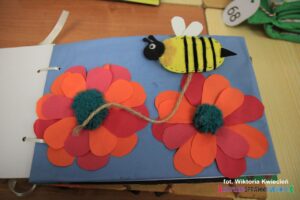Wnętrze książki dotykowej w zółto-czarne paski pt. Mała Wielka Pszczoła; na jednym z dwóch czerwonych kwiatów "przysiadła" pszczoła wykonana z filcu