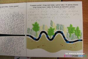Wnętrze książki pt. "Moja droga" z dotykową ilustracją i napisem w języku Braille'a oraz polskim: "Przestało padać. Droga jest kręta i pełna kałuż. Po jednej stronie drogi rosną trawy i ziela. Po drugiej są kamienie i piasek."