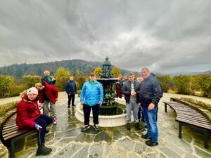 Grupa Uczestników zgromadzona wokół fontanny w Ogrodach Zmysłów w Muszynie; w tle panorama na góry