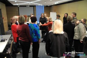 Pani Ewa Jasińska, prowadząca warsztat pt. "Jak zbudować skuteczny zespół" tłumaczy zebranym wokół uczestnikom sposób wykonania ćwiczenia