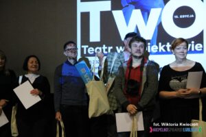 Uczestnicy konkursu stoją w rzędzie, w tle ekran multimedialny z wyświetlonym plakatem TwO