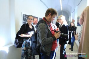 Na pierwszym planie mężczyzna z aparatem fotograficznym, w tle ludzie zgromadzeni na wernisażu