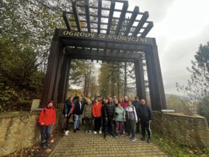 Grupa uczestników stojąca pod drewnianą, geometryczną konstrukcją z napisem "OGRODY SENSORYCZNE"