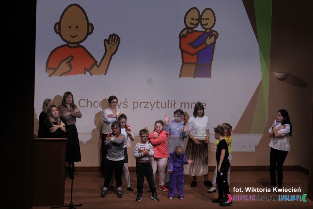 Na scenie stoi grupa dzieci wraz z instruktorami, wykonująca gest obejmowania się rękami, w tle na ekranie multimedialnym wyświetlony jest slajd z obrazkiem postaci i napisem "Chcę byś przytulił mnie"