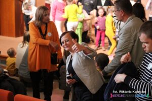 Kobieta unosi kciuk w górę w geście aprobaty, uśmiecha się do obiektywu, w tle na scenie dużo osób