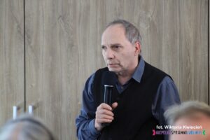 Na tle drewnianych drzwi stoi mężczyzna w granatowej koszuli i ciemnej marynarce, w ręku trzyma mikrofon