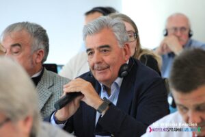 Pomiędzy innymi uczestnikami siedzi mężczyzna w granatowej marynarce, w ręku trzyma mikrofon