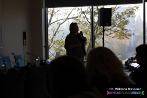 Zacieniona sylwetka kobiety (dr hab. Anny Prokopiak), stojącej na tle jesiennego drzewa, widocznego za oknem w blasku światła