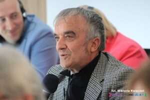 Mężczyzna w marynarce w drobną kratkę mówi do trzymanego przez siebie mikrofonu