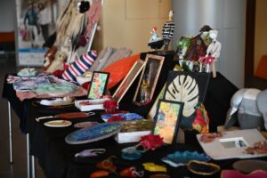 Stół ekspozycyjny z różnymi rękodziełami, w tym obrazami, maskami, przedmiotami dekoracyjnymi, wszystkie ułożone na czarnym obrusie.