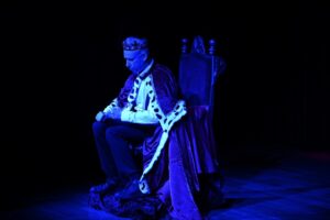 Mężczyzna ubrany jak król siedzi na tronie. Nosi koronę i królewską szatę z futrzanym wykończeniem, otoczona przyćmionym światłem, które rzuca niebieski odcień na scenę.