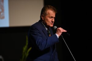 Mężczyzna w niebieskim garniturze mówi do mikrofonu. W tle widoczny częściowo ekran