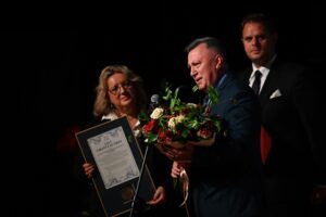 Mężczyzna w garniturze trzyma bukiet kwiatów i wygłasza przemówienie do mikrofonu. U jego boku stoi kobieta trzymająca oprawiony list gratulacyjny. W tle widoczny jest inny mężczyzna w garniturze.