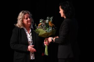 Kobieta o jasnych włosach sięga po bukiet kwiatów od drugiej brunetki, która jej podaje. Obie są ubrane w czarne garnitury.