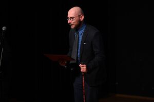 Mężczyzna w garniturze i krawacie stoi na scenie i trzyma w jednej ręce kij nordic walking, a w drugiej dyplom. Zmierza do mikrofonu.