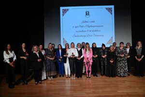 Grupa kobiet i jeden mężczyzna stoją na scenie przed dużym ekranem z napisem: Kultura i sztuka w życiu osób z niepełnosprawnościami na przestrzeni 15 lat działalności Środowiskowego Domu Samopomocy. Wiele kobiet trzyma róże.