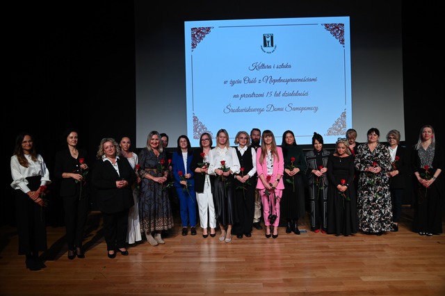 Grupa kobiet i jeden mężczyzna stoją na scenie przed dużym ekranem z napisem: Kultura i sztuka w życiu osób z niepełnosprawnościami na przestrzeni 15 lat działalności Środowiskowego Domu Samopomocy. Wiele kobiet trzyma róże.