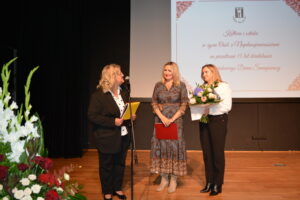 Trzy kobiety stoją na scenie. Jedna mówi do mikrofonu, druga trzyma czerwoną teczkę, a trzecia trzyma bukiet kwiatów. W tle na ekranie widnieje tytuł wydarzenia: Kultura i sztuka w życiu osób z niepełnosprawnościami na przestrzeni 15 lat działalności Środowiskowego Domu Samopomocy.