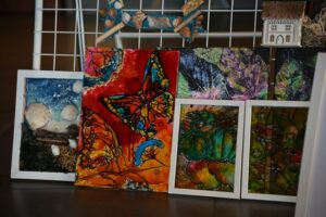 Kolorowe dzieła sztuki przedstawiające m.in. motyle, muszle i elementy o tematyce liści, prezentowane na drucianej siatce