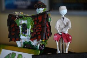 Ręcznie wykonana figurka ubrana w materiałowy strój siedzi na stołku, obok miniaturowego domu z teksturowanym dachem i zielenią. Dom ma małą latarnię wiszącą na sznurku