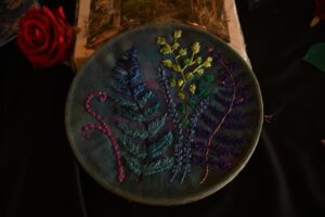 Okrągła podstawka z materiału haftowana kolorowymi wzorami paproci i roślin w odcieniach niebieskiego, fioletu, żółtego i zieleni, umieszczonymi na ciemnym tle. Obok podstawki czerwona róża.