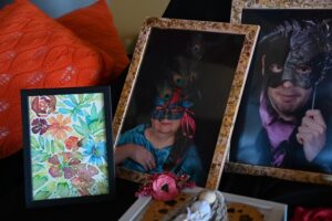 Dwa oprawione zdjęcia kobiety i mężczyzny w maskach. Obok zdjęć stoi ramka z obrazem przedstawiającym kolorowe kwiaty i pomarańczowe poduszki