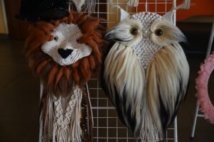 Dwa dzieła sztuki makramy wystawione na drucianej siatce: jedno przedstawiające lwa z brązową grzywą, a drugie sowę z dużymi oczami i teksturowanym ciałem.