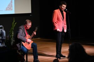 Mężczyzna w czerwonej marynarce stoi na scenie przy mikrofonie i śpiewa. Obok niego siedzi inny mężczyzna i gra na gitarze. Występują pod światłami scenicznymi, z dużym ekranem w tle.