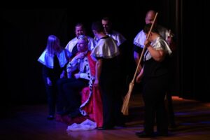 Grupa ludzi w ciemnych ubraniach i białych pelerynkach otacza siedzącą osobę ubraną w koronę i czerwoną pelerynę. Obok stoi mężczyzna trzymający miotłę.