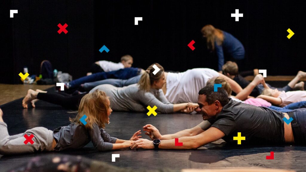 Grupa dorosłych i dzieci leży na podłodze, sięgając rękami do siebie.