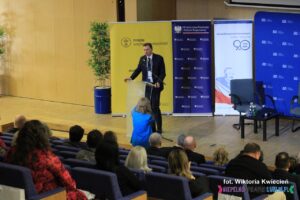 Na scenie za pulpitem stoi Pan Mariusz Jedynak, Członek Zarządu nadzorującego Pion Administracji i Zamówień Publicznych; widok z widowni