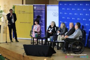 Na scenie stoi mężczyzna z mikrofonem - Pan Mariusz Jedynak, siedzący uczestnicy drugiego panelu patrzą w Jego stronę