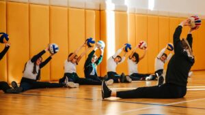 Grupa dziewczynek siedzi na podłodze sali gimnastycznej, unosząc piłki do siatkówki nad głowami, podczas ćwiczeń prowadzonych przez instruktorkę.