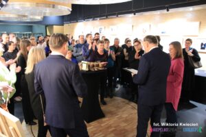 Oficjalne otwarcie wernisażu; grupa ludzi zgromadzona w oświetlonej sali, na ścianach wisi rząd obrazów