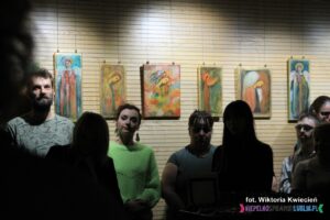 Pod ścianą w rzędzie stoją uczestnicy wernisażu, na ścianie nad nimi wiszą kolorowe obrazy z Aniołami