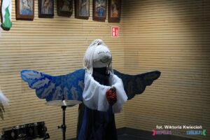 Figura uśmiechniętego Anioła; na ścianie powyżej widać dolną część wiszących w rzędzie obrazów