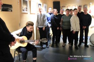 Na pierwszym planie siedzi kobieta grająca na gitarze, w tle stoi grupa osób tworzących "Anielski Chór"