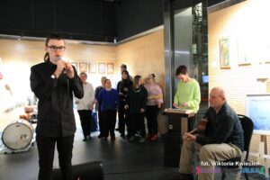 Z mikrofonem stoi solista Kajetan Rachwał, obok siedzi mężczyzna z gitarą, nieco z tyłu kobieta przy cymbałkach a w tle grupa osób tworzących "Anielski Chór"