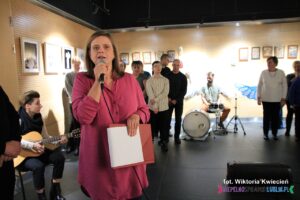 Z mikrofonem stoi Pani Agnieszka Błaszczak, Kierownik Środowiskowego Domu Samopomocy „Absolwent”, w tle za Nią stoją wykonawcy "Anielskiego Chóru"