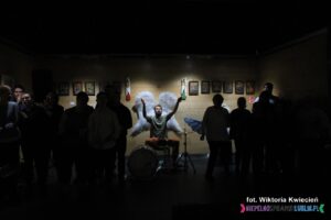 Centralnie w najlepiej oświetlonym punkcie, przy perkusji siedzi mężczyzna z uniesionymi dłońmi, za jego plecami na ścianie wiszą białe skrzydła, z przodu w mroku widać sylwetki stojących osób