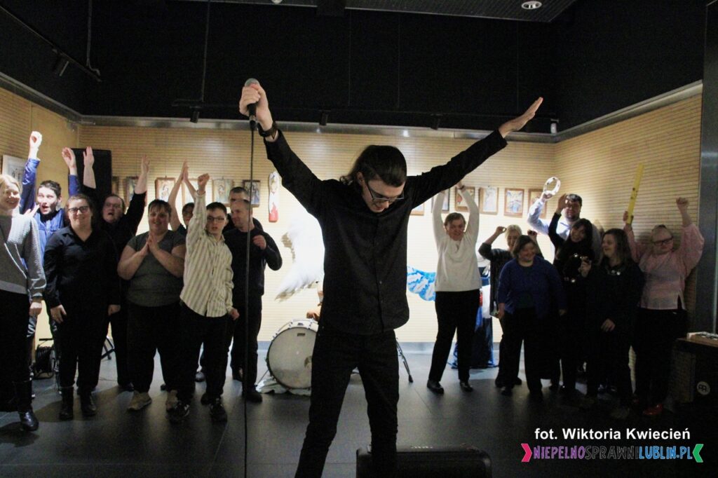 Solista unosi ręce w górę, w tle po jego obu stronach grupy osób unoszą ręce, są radośni