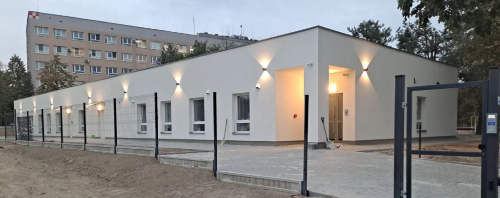 Parterowy, biały budynek z zewnętrznym oświetleniem, otoczony płotem, położony przed wielopiętrowym blokiem mieszkalnym.
