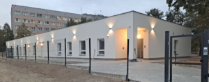 Parterowy, biały budynek z zewnętrznym oświetleniem, otoczony płotem, położony przed wielopiętrowym blokiem mieszkalnym.