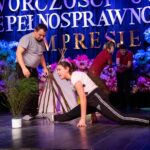 Na pierwszym planie widoczna jest kobieta wykonująca szpagat. W tle znajdują się cztery osoby, które wykonują różne ćwiczenia; jeden z mężczyzn podnosi hantle. Obok kobiety robiącej szpagat stoi mała zielona roślina doniczkowa.